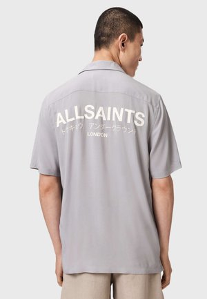 Mann trägt ein graues Kurzarmshirt mit weißem Schriftzug „ALLSAINTS Tokyo Underground London“ auf dem Rücken und steht vor einem einfarbigen Hintergrund.
