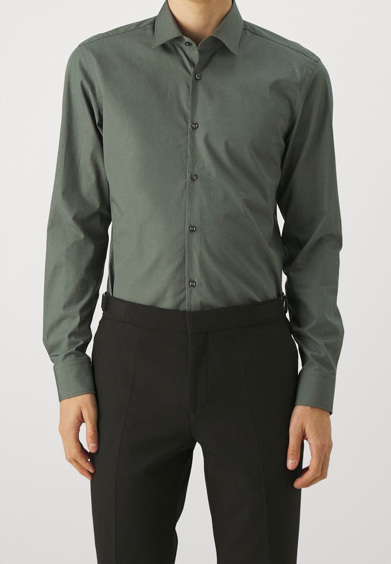 Homme portant une chemise ajustée vert olive à boutons avec un pantalon de costume noir, les mains détendues le long du corps, debout devant un fond uni.