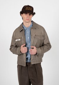 Unge mand iført brun kasket med teksten "The Art of Being Late", brun jakke, blå skjorte med striber og brune bukser, poserer foran en ensfarvet hvid baggrund.