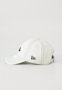 Fehér pamut baseball sapka ívelt vízszinttel, fekete hímzett logóval elöl, és fekete kiegészítő logóval az oldalon. Öltögetett részletekkel rendelkezik.
