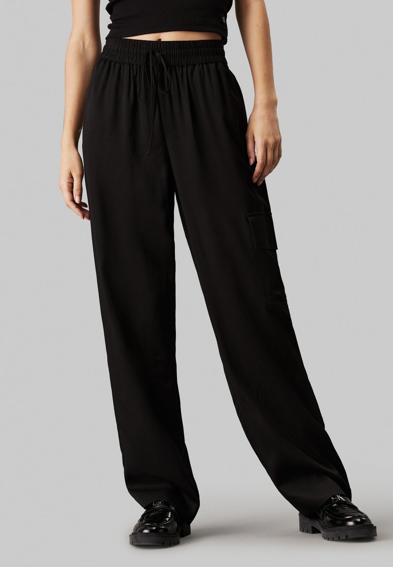 Sorte, brede bukser lavet af et glat materiale, med en elastisk talje med snor og en sidelomme.