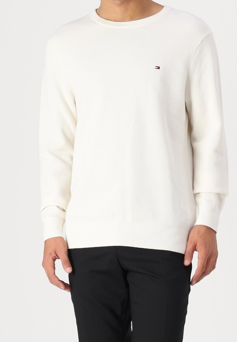 Homme portant un pull blanc uni à manches longues avec un petit logo rouge, blanc et bleu sur la poitrine et un pantalon noir sur fond blanc.