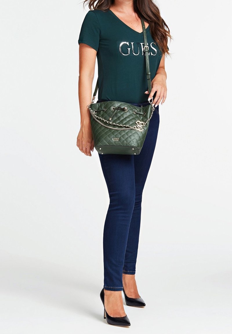 Femme portant un t-shirt Guess vert foncé et un jean bleu, tenant un sac à bandoulière matelassé vert avec un détail en chaîne et des talons hauts noirs.