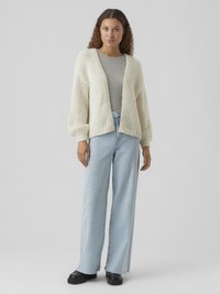 Donna in piedi che indossa un cardigan bianco lavorato a maglia spesso, una t-shirt grigia, jeans larghi azzurri e scarpe nere su uno sfondo grigio uniforme.
