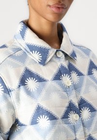 Chaqueta azul con patrón que presenta diseños triangulares, motivos de ráfaga blanca, acentos en color beige claro, un cuello y botones. Primer plano de la tela texturizada.