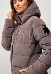 Chaqueta acolchada en marrón apagado, con un alto collar, cierre de cremallera y un parche negro de copo de nieve en la manga. Textura suave y acolchada.