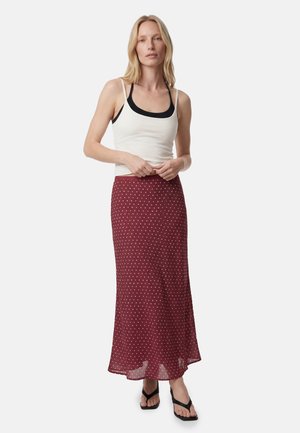 Blondynka ubrana w biały tank top na czarnym sportowym staniku, długą bordową spódnicę w białe kropki i czarne klapki.