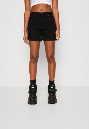 Person iført sorte shorts med høj talje og op-rullet kant, sorte sokker og chunky sorte sneakers, stående mod en ensfarvet baggrund.