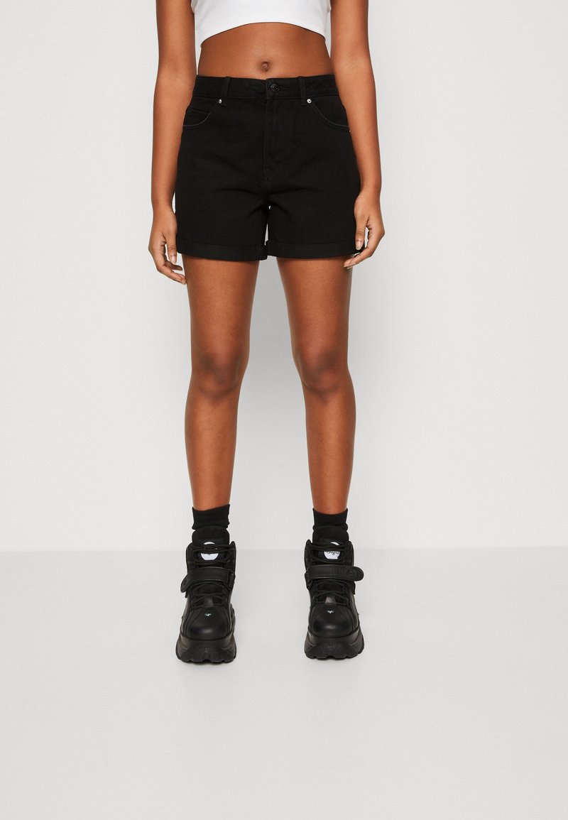 Personne portant un short noir taille haute avec ourlet retroussé, des chaussettes noires et des baskets noires épaisses, debout devant un fond uni.