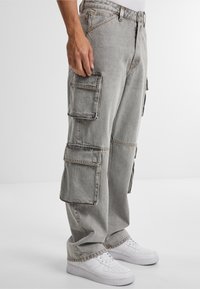 Pantaloni cargo grigi con multiple tasche laterali e cuciture visibili, abbinati a sneakers bianche. Il tessuto sembra resistente, con una vestibilità comoda.