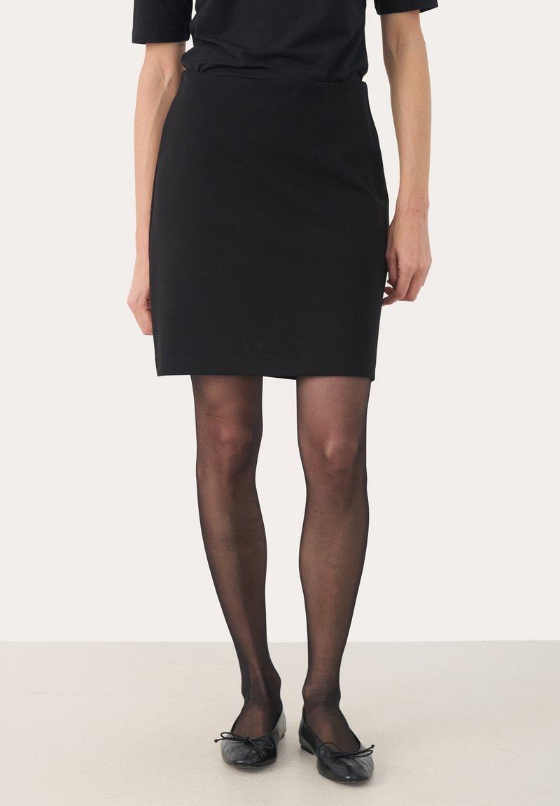 Femme portant une jupe noire longueur genou, des collants noirs transparents, des ballerines noires et un haut noir à manches courtes sur un fond uni.