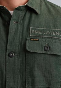 Chemise verte à motif en chevrons, avec des boutons noirs, une poche poitrine et une marque "PME Legend" brodée sur la poche et le tissu.