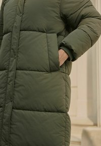 Geacă puf verde cu un design matlasat, având un buzunar lateral, manșete cu bordură și închidere cu capse în față. Țesătură netedă și texturată.