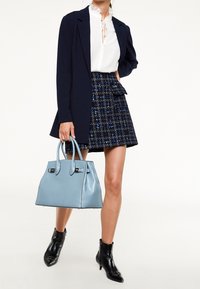 Ljusblå handväska med strukturerad design och dubbla handtag. Modellen bär en vit blus med volangkrage, marinblå jacka och mönstrad kjol. Svarta ankelboots.