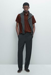 Polo shirt marrone, maglione grigio drappeggiato sulle spalle, pantaloni grigio scuro e scarpe marroni; tessuto morbido e vestibilità comoda.
