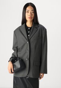 Gråstripete blazer i strukturert design med knapper og en brystlomme. Modellen holder en liten svart lærveske og har på seg en mørk topp.