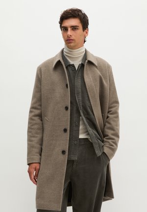 Jeune homme portant un pardessus beige boutonné, une veste en velours côtelé grise, un col roulé crème et un pantalon en velours côtelé gris assorti, debout avec une main dans la poche.
