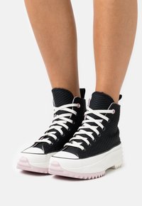 Svarta högskurna sneakers med textilerad tyg, vita snören och en kontrasterande rosa gummisula med tandad kant.