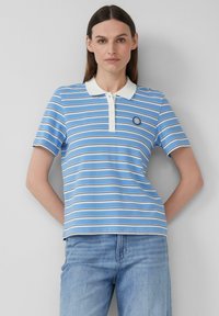 Frau trägt ein blau-weiß gestreiftes Poloshirt mit weißem Kragen und hellblauen Jeans, steht mit den Händen hinter dem Rücken.