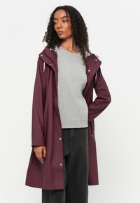 Casaco impermeável bordô com capuz, fechos de pressão e cordão. Usado sobre um moletom cinza claro e jeans escuros, apresentando um tecido texturizado.