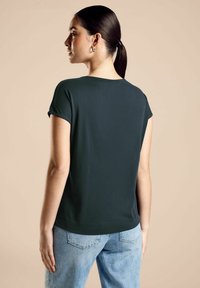 Maglietta a maniche corte verde scuro con scollatura rotonda, realizzata in tessuto morbido. Abbinata a jeans in denim azzurro, che mostrano un fitting informale.