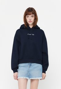 Mørkeblå hettejakke med snøring og ribbestrikkede mansjetter. Har brodert "Tommy Hilfiger"-logo på forsiden. Kombinert med et slitt denimskjørt.
