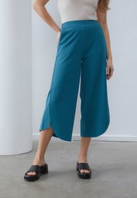 Teal vide bukser med sideslids, der har en glat tekstur og mid-rise talje. Båret med sorte platform sandaler.