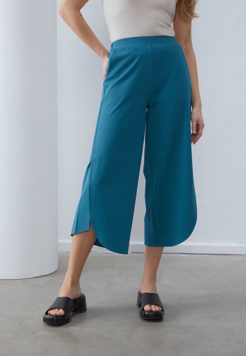 Teal vide bukser med sideslids, der har en glat tekstur og mid-rise talje. Båret med sorte platform sandaler.