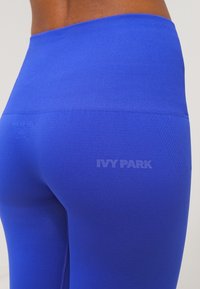 Vysoké modré legíny z hladkého, elastického materiálu. Obsahují embosované logo "IVY PARK" blízko pasu a elegantní texturované detaily.