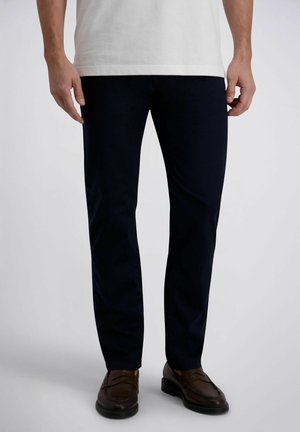 Mężczyzna w ciemnogranatowych spodniach, brązowych skórzanych mokasynach i białej koszuli z krótkim rękawem stoi na jednolitym tle.