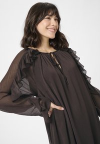 Nasmejana žena s kestenjastom kosom do ramena, odjevena u labavu, prozirnu tamnosmeđu haljinu s naboranim rukavima i vratom s mašnom.