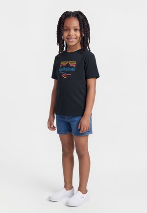 Παιδί με πλεξούδες, φορώντας μαύρο μπλουζάκι Quiksilver, τζιν σορτς και λευκά παπούτσια slip-on, στέκεται και χαμογελά σε λευκό φόντο.