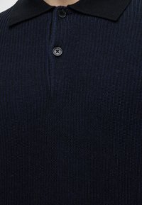 Gros plan sur un polo en maille côtelée bleu marine foncé avec col noir et deux boutons, porté par une personne.