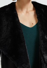 Černý límec z umělé kožešiny s pod ním zeleným žebrovaným svetrem. Detailní pohled, který ukazuje kontrast textur a tvar výstřihu.