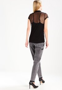 Camiseta negra con espalda de malla transparente y estampada, y mangas cortas, combinada con jeans gris ajustados y tacones altos negros.