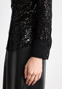 Schwarze Paillettenjacke mit einer strukturierten Oberseite, glattem Stoff unten und geknöpften Bündchen. Bemerkenswerte Akzente sind runde Knöpfe an den Bündchen.