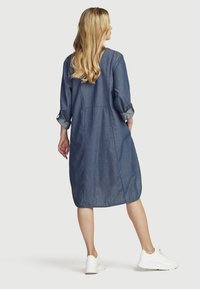 Denim shirtjurk met lange mouwen, een losse pasvorm en een lengte tot halverwege de kuiten. Heeft opgerolde manchetten en een afgeronde zoom; gedragen met witte sneakers.