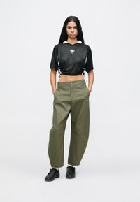 Svart croppad mesh-topp med vit krage och sidosnören, kombinerad med olivgröna vida byxor. Svarta sportskor kompletterar outfiten.