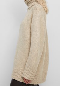 Beige värinen pooloneulepusero, valmistettu paksusta neuloskankaasta, jossa on resorit hihansuissa ja helmassa. Tarjoaa väljähkön istuvuuden ja tekstuurisen ulkonäön.