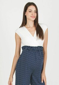 Combinaison bleu marine à pois blancs, avec un haut enroulé blanc et une taille smockée pour une silhouette ajustée. Texture de tissu douce.