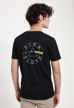 Młody mężczyzna zwrócony tyłem, ubrany w czarną koszulkę z motywem pryzmatu Pink Floyd i tęczowym światłem na plecach, beżowe spodnie, kręcone ciemne włosy.