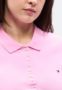 Γυναίκα που φοράει ένα ανοιχτό ροζ πόλο πουκάμισο με τέσσερα κουμπιά και ένα μικρό λογότυπο Tommy Hilfiger στο στήθος, εμφανιζόμενη από το πηγούνι έως τον άνω κορμό.