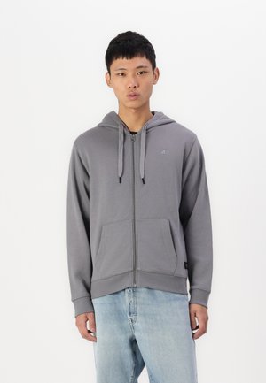 Grauer Reißverschluss-Hoodie mit einer Kordelzugkapuze, Fronttaschen und einem kleinen Logo auf der Brust. Weicher Stoff und lockere Passform. Kombiniert mit hellblauen Jeans.