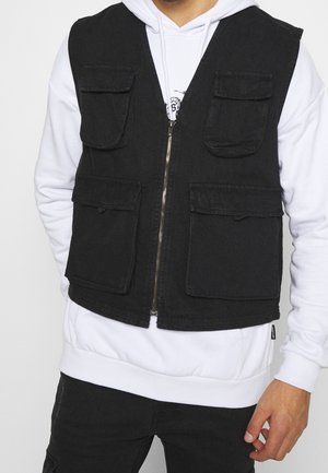 Homme portant un sweat à capuche blanc sous un gilet sans manches noir avec quatre poches à rabat à l'avant et une fermeture éclair frontale.