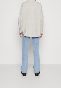 Person wearing loose light blue jeans, black shoes, and a long-sleeve beige shirt with thin vertical stripes, standing against a white background.