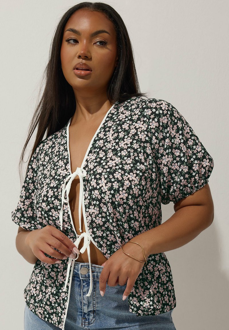 Blommig kortärmad blus med djup V-halsringning och knytning, med svart bakgrund, rosa blommor och ett lätt tyg.