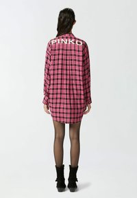 Camicia oversize a quadri rosa e nera con maniche lunghe, caratterizzata da un logo bianco "PINKO" sul retro. Abbinata a stivali in suede neri.