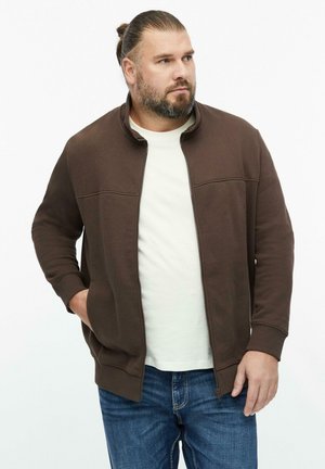 Mann mit Bart und gebundenem Haar, trägt eine braune Jacke mit Reißverschluss, ein weißes Hemd und blaue Jeans, steht mit einer Hand in der Tasche.