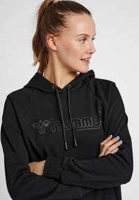 Czarna bluza z kapturem z logo na przodzie, sznurkami do regulacji i rękawami raglanowymi. Miękka faktura, luźny krój, bez widocznych wzorów czy akcentów.