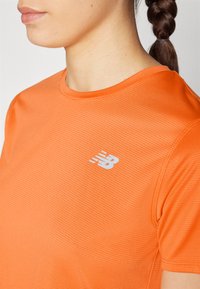 Oranžna športna majica iz teksturiranega materiala. Ima okroglo ovratnico in belo New Balance logo na levi strani prsnega koša.